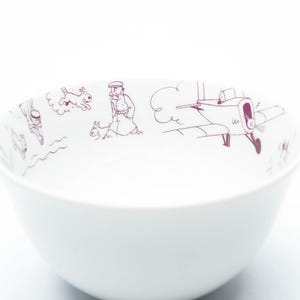 May include: White ceramic bowl with a purple illustration around the rim. The illustration depicts a person, dogs, and an airplane. The bowl has a simple, rounded shape and is set against a white background.