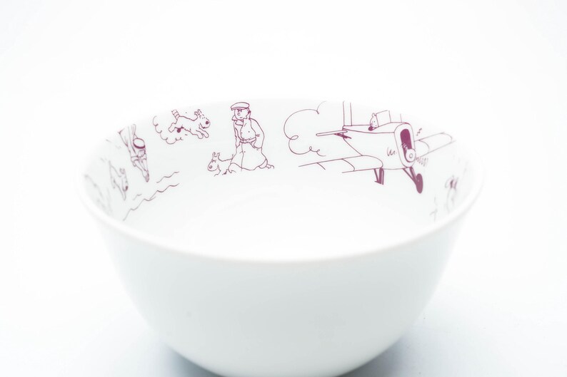 May include: White ceramic bowl with a purple illustration around the rim. The illustration depicts a person, dogs, and an airplane. The bowl has a simple, rounded shape and is set against a white background.