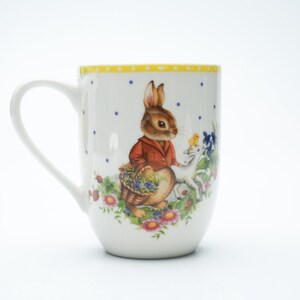 Puede incluir: Taza de cerámica blanca con una ilustración de un conejo con chaqueta roja que lleva una cesta, con un cervatillo y flores. El borde tiene un diseño de lunares amarillos. La taza tiene un asa curva.