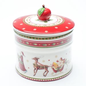 Op de afbeelding: Een keramische kerstkoekjespot met een rood deksel en een appelvormige handgreep. De pot heeft feestelijke illustraties van de kerstman, rendieren en een slee. De pot is wit met rode en groene accenten.