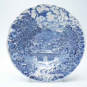 Puede incluir: Un plato redondo de cerámica con un patrón de transferencia azul y blanco. El diseño muestra un paisaje pastoral con un puente de piedra sobre un arroyo, árboles grandes, una iglesia a lo lejos y una persona pescando en la orilla.