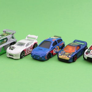 May include: Six toy cars lined up on a green background. The cars are all different colours and designs, including a white car with red and black flames, a blue car with a white number 12, a white car with a green and silver police design, and a white car with a red and black design.
