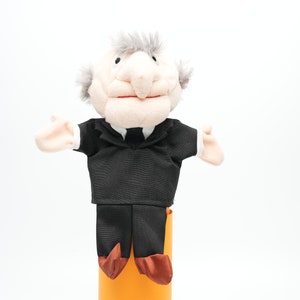 May include: A plush puppet of an older person wearing a black suit and a white collared shirt. The puppet has grey hair and a big smile.