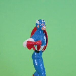 K&ouml;nnte beinhalten: Eine Actionfigur von Captain America, die einen blauen und wei&szlig;en Anzug mit einem roten Umhang tr&auml;gt und einen rot-wei&szlig;-blauen Schild h&auml;lt. Die Figur steht auf einem gr&uuml;nen Hintergrund.