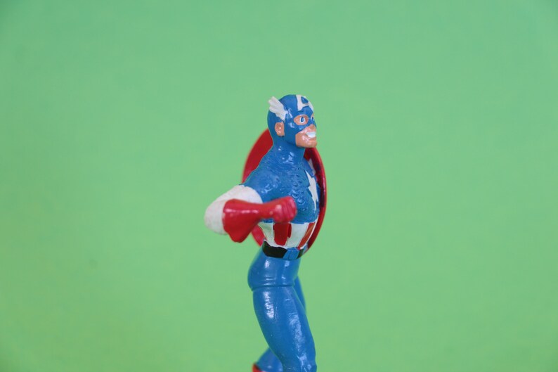 K&ouml;nnte beinhalten: Eine Actionfigur von Captain America, die einen blauen und wei&szlig;en Anzug mit einem roten Umhang tr&auml;gt und einen rot-wei&szlig;-blauen Schild h&auml;lt. Die Figur steht auf einem gr&uuml;nen Hintergrund.