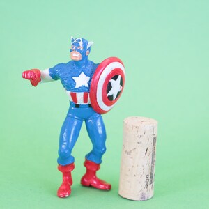 K&ouml;nnte beinhalten: Eine Plastik-Actionfigur von Captain America, die mit erhobenem Schild steht. Er tr&auml;gt ein blaues und rotes Kost&uuml;m mit einem wei&szlig;en Stern auf der Brust. Er steht neben einem braunen Korken.
