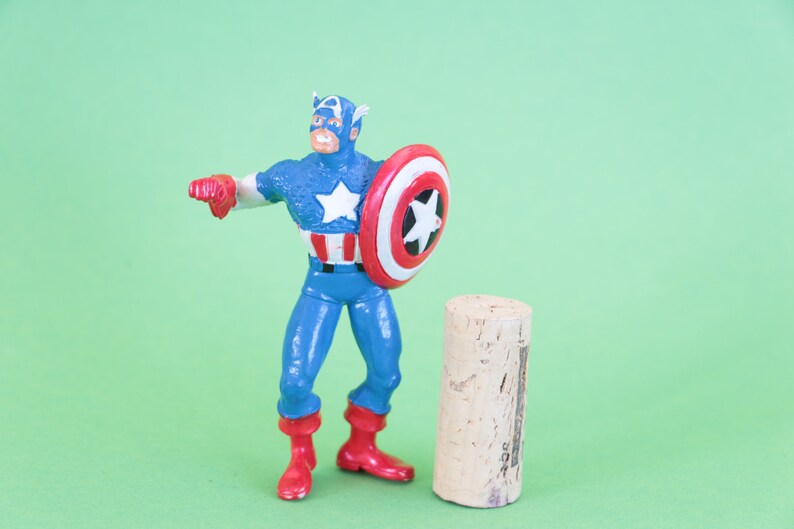 K&ouml;nnte beinhalten: Eine Plastik-Actionfigur von Captain America, die mit erhobenem Schild steht. Er tr&auml;gt ein blaues und rotes Kost&uuml;m mit einem wei&szlig;en Stern auf der Brust. Er steht neben einem braunen Korken.