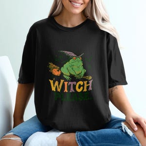 May include: A black short-sleeve t-shirt featuring a graphic of a green frog wearing a purple witch hat and riding a broomstick with an orange pumpkin. Below the frog, the text reads WITCH PLEASE in large, colorful, retro-style lettering.