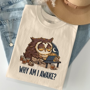 May include: A cream-colored t-shirt featuring a graphic of a tired brown owl sitting at a small desk with a laptop and several coffee cups. The text below the illustration reads, "WHY AM I AWAKE?" The shirt is displayed alongside blue denim jeans and a tan hat.
