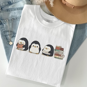 May include: White t-shirt with a graphic of three cartoon penguins. One reads a book, another stands, and the third rests near a stack of books. The shirt is laid on blue denim jeans.