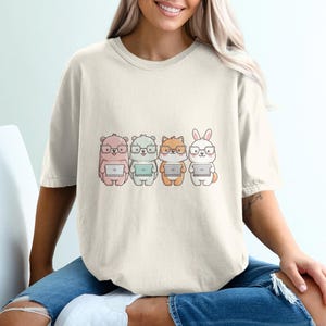 Puede incluir: Una camiseta de manga corta de color crema con un estampado gráfico de cuatro animales de dibujos animados de pie en fila. Cada animal lleva gafas y sostiene un pequeño ordenador portátil. Los personajes incluyen un oso rosa, un oso blanco, un zorro marrón y un conejo blanco.