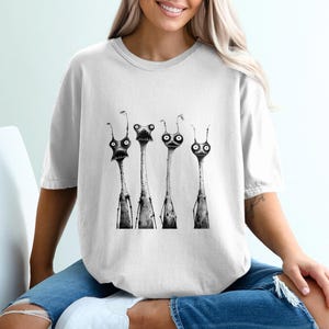 May include: White t-shirt featuring a black and white illustration of four elongated creatures with large eyes and detailed features. The creatures have thin bodies and are arranged vertically. The shirt is a comfortable fit.