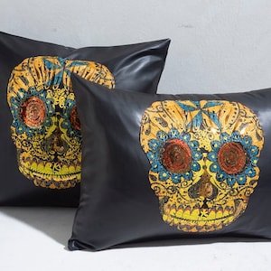 Puede incluir: Dos cojines decorativos negros con un diseño de calavera colorido. Las calaveras presentan patrones florales amarillos, azules y naranjas. Los cojines tienen una textura suave similar al cuero.