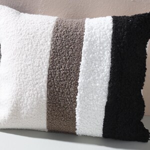Pode incluir: Uma almofada decorativa retangular com um design listrado texturizado. A almofada apresenta listras verticais em branco, marrom e preto. O tecido tem uma aparência macia e felpuda, adequada para decoração de casa.
