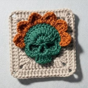 Puede incluir: Un cuadrado de ganchillo con un diseño de calavera verde y pétalos de flores naranjas. La calavera está centrada en un cuadrado beige, mostrando un aspecto hecho a mano y texturizado. Probablemente un diseño de parche o aplique.