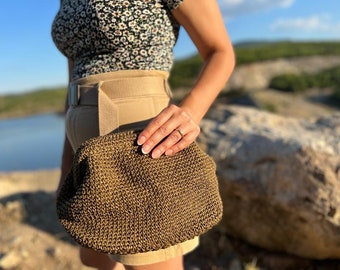 May include: A woven, olive-green clutch bag with a rounded shape. The bag is held by a person wearing a floral top and tan shorts. The background features a lake and rocky terrain.