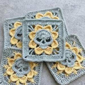 Puede incluir: Cinco cuadrados de ganchillo con un motivo de calavera. Cada cuadrado presenta un diseño de calavera en el centro, rodeado de pétalos de flores amarillas, sobre un fondo azul claro. Los cuadrados están hechos a mano y tienen una apariencia texturizada.