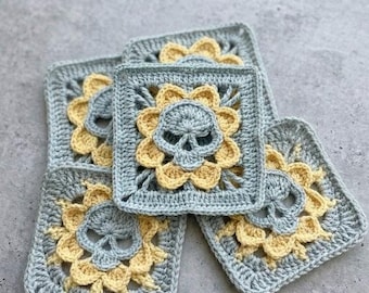May include: A collection of five crocheted squares. Each square features a skull design in the center, surrounded by yellow flower petals, all set against a light blue background. The squares are handmade and have a textured appearance.