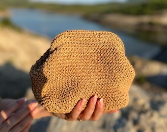 Puede incluir: Un bolso de mano tejido de color marrón claro con una superficie texturizada. El bolso se sostiene contra un fondo borroso de agua y rocas. El bolso de mano tiene una forma redondeada y una textura tejida.