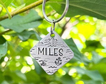 Puede incluir: Llavero de plata con una etiqueta en forma de diamante que dice "MILES" con un diseño de olas y un ancla. La etiqueta está colgada de un llavero de plata.