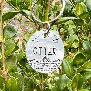 May include: Silver keychain with a round charm featuring a stylized ocean scene with waves and seagulls. The charm has the word "OTTER" engraved on it.