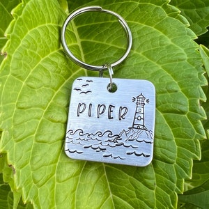 May include: Silver keychain with a square tag featuring a lighthouse and waves. The tag is engraved with the name "PIPER".