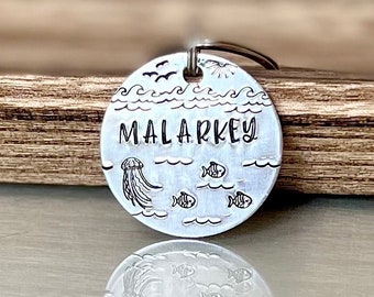 May include: A circular, silver metal tag with the word "MALARKEY" etched in the centre. The tag features etched images of waves, fish, a jellyfish, and birds. A small metal ring is attached at the top.