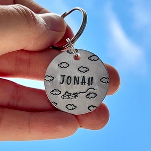 May include: Silver keychain with a round tag featuring a plane flying through clouds and the name "JONAH" engraved on it.