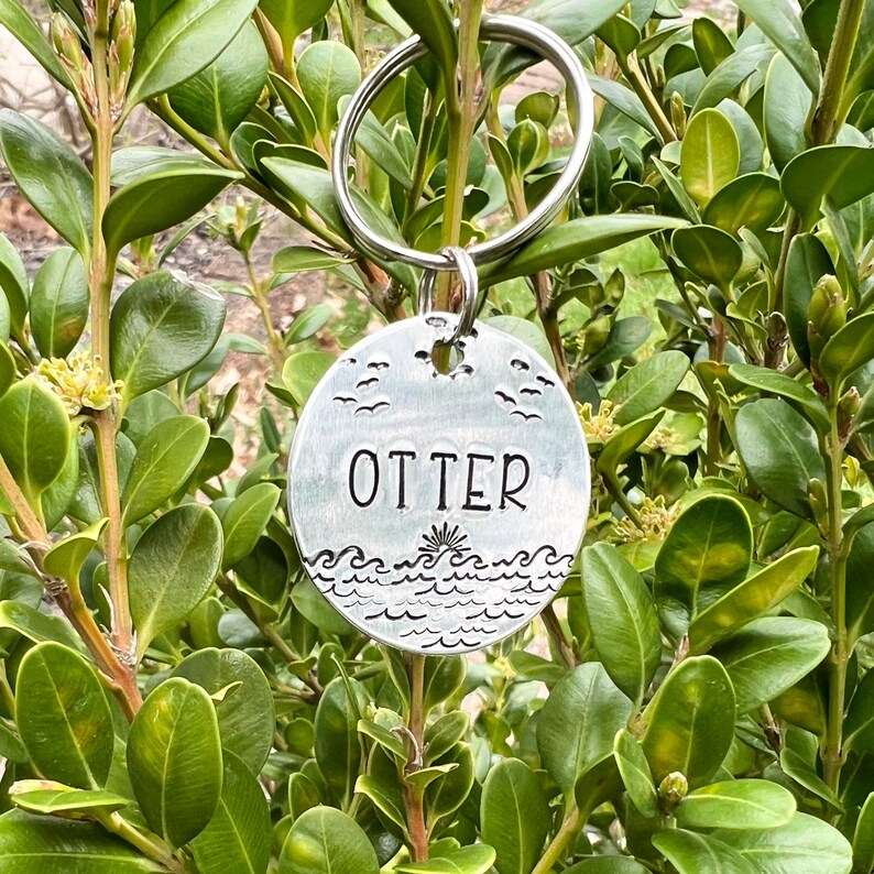 May include: Silver keychain with a round charm featuring a stylized ocean scene with waves and seagulls. The charm has the word "OTTER" engraved on it.