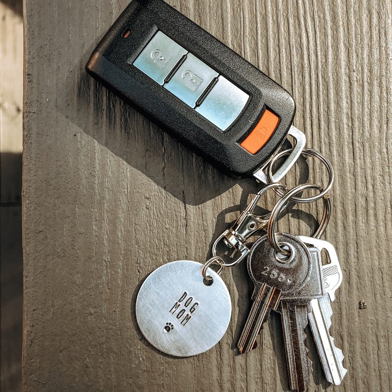 May include: A silver key chain with a round metal tag that says "Dog Mom" and a paw print. The keychain also has a black car key fob and three silver keys.