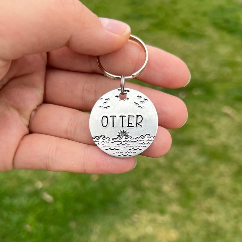 May include: Silver key chain with a round tag that says "OTTER" and has a design of waves and birds.