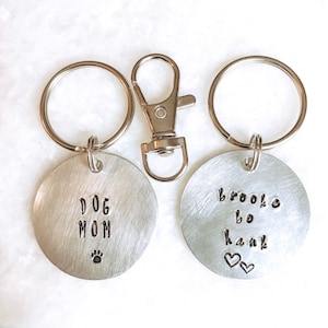 May include: Two silver keychains with round metal tags. One tag says "Dog Mom" with a paw print. The other tag says "Brooke bo Hank" with two hearts.