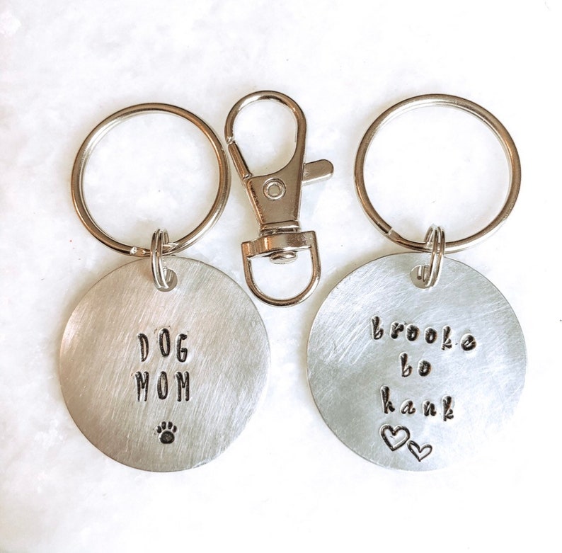 May include: Two silver keychains with round metal tags. One tag says "Dog Mom" with a paw print. The other tag says "Brooke bo Hank" with two hearts.