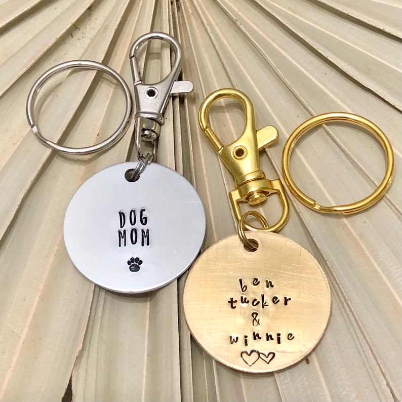 May include: Two silver and gold keychains with round metal tags. The silver tag says "Dog Mom" with a paw print. The gold tag says "Ben Tucker & Winnie" with two hearts.