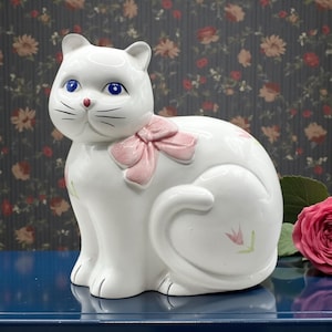 Könnte beinhalten: Eine weiße Keramik-Katzenfigur mit blauen Augen, einer rosa Schleife und rosa Blumenakzenten. Die Katze sitzt aufrecht mit eingerolltem Schwanz. Die Figur steht auf einer blauen Oberfläche mit floralem Hintergrund.