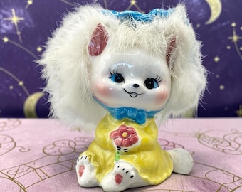 May include: A ceramic figurine of a white animal with large blue eyes, pink cheeks, and long white faux fur trim around its head. The figure wears a yellow dress with a blue collar and holds a small pink flower in its hands, with pink paw pads.