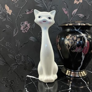 May include: A white ceramic cat figurine with a long neck and a whimsical expression. The cat has pink accents on its cheeks and mouth. Beside it is a black urn with gold floral designs.