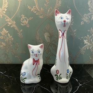 May include: Two white ceramic cat figurines with hand-painted details. The shorter cat sits, while the taller cat stands. Both feature blue eyes, pink ears, a red nose, and a pink bow around the neck, accented with small blue and red floral motifs.