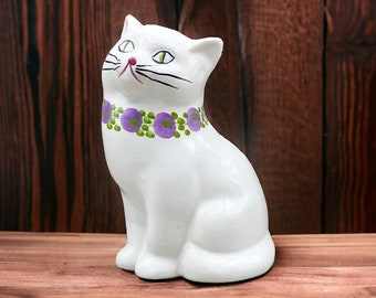 Könnte beinhalten: Weiße Keramik-Katzenfigur mit violetten und grünen Blumendetails am Hals. Die Katze sitzt mit erhobenem Kopf.