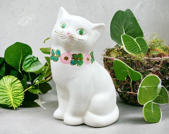 Könnte beinhalten: Weiße Keramik-Katzenfigur mit grünen und rosa Blumendetails um den Hals. Die Katze sitzt mit dem Kopf nach oben geneigt und hat grüne Augen.