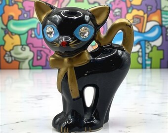 May include: A black ceramic cat figurine with gold accents on the ears, tail, and bow tie. The cat has blue eyes with rhinestone accents and a red nose. The figurine is standing upright.