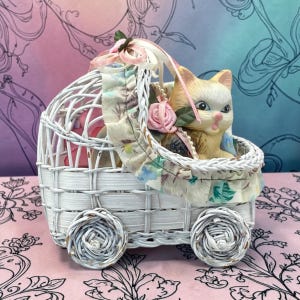 Op de afbeelding: Een wit rieten kinderwagenfiguurtje met een klein keramieken kitten met crème en beige vacht. De wagen is versierd met een bloemrijke stoffen rand, roze lintstrikjes en een klein roze stoffen roosje, rustend op vier geweven rieten wielen.