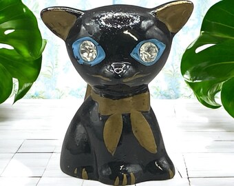 May include: A black ceramic cat figurine with gold accents on the ears and bow tie. The cat has large, sparkling blue eyes. The figurine is a decorative item, likely for a shelf or tabletop.