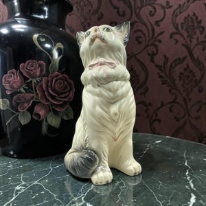 Könnte beinhalten: Eine weiße Keramik-Katzenfigur mit rosa Schleife, die nach oben blickt. Die Katze hat grüne Augen und einen flauschigen Schwanz mit grauen Akzenten. Die Figur steht auf einer marmorähnlichen Oberfläche, im Hintergrund eine schwarze Vase mit Rosendetails.