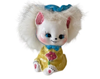 May include: A white ceramic figurine of a cat wearing a blue hat and a yellow dress. The cat has pink ears, blue eyes, and a pink nose. It is holding a pink flower.