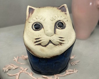 May include: A ceramic cat figurine with a cream-colored face and dark blue body. The cat has large, detailed eyes and a smiling expression. The figurine is decorated with brown accents and sits on a gray surface with pink yarn embellishments.