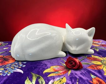 May include: A smooth, white ceramic figurine of a cat in a curled, sleeping position. The cat has a glossy finish and is positioned next to a single, small, deep red rose bud. The sculpture features simple, clean lines and a rounded, compact form.