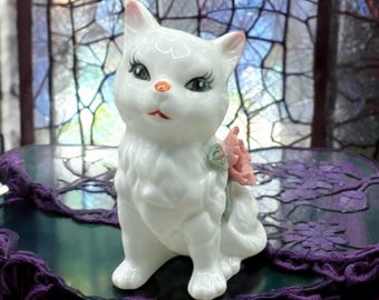 May include: A white porcelain figurine of a cat with pink flowers. The cat is sitting with its head tilted to the side and has green eyes. The figurine is detailed with a white fur texture and a pink flower on its shoulder.