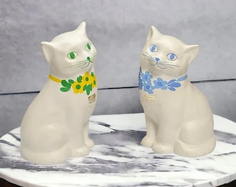 Könnte beinhalten: Zwei weiße Keramik-Katzenfiguren mit bemalten Details. Eine Katze trägt eine grüne und gelbe Blumengirlande, die andere eine blaue Blumengirlande.