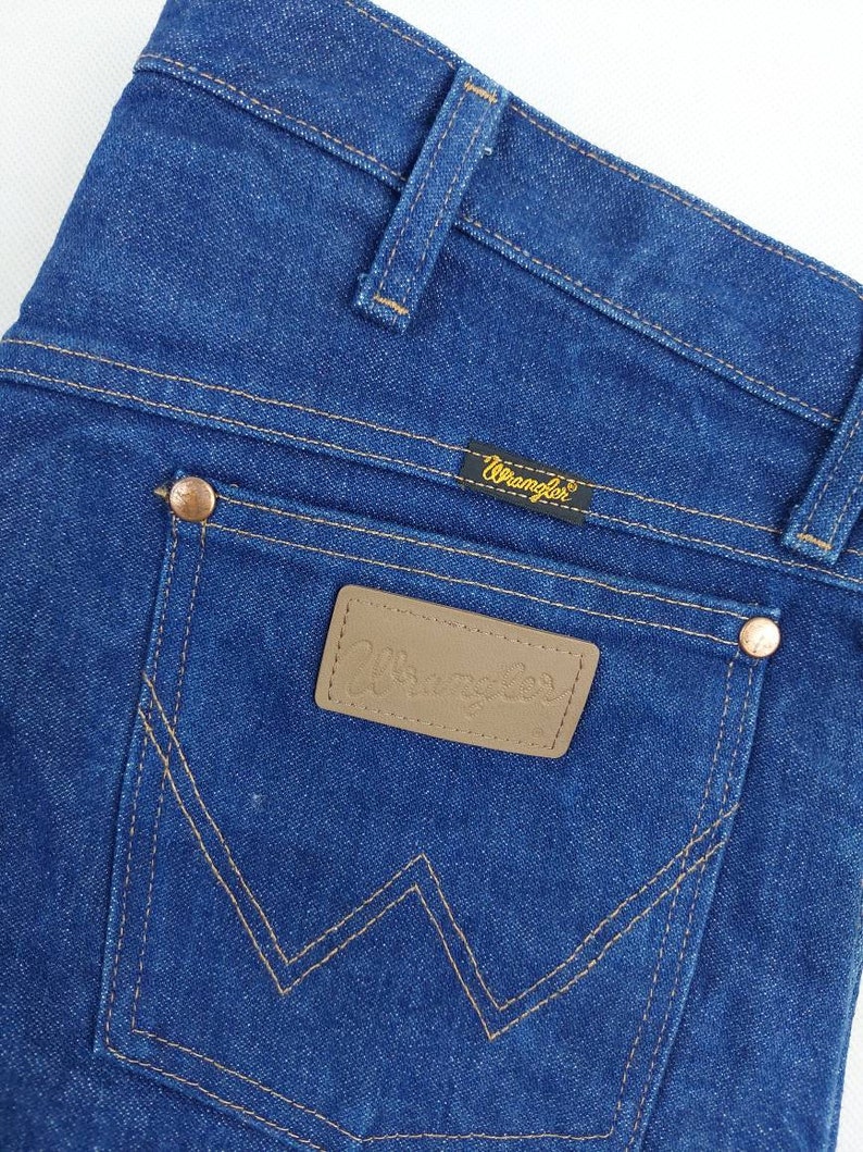 May include: A pair of blue denim jeans with a brown leather patch that says "Wrangler" on it. The jeans have a classic five-pocket style and a straight leg.
